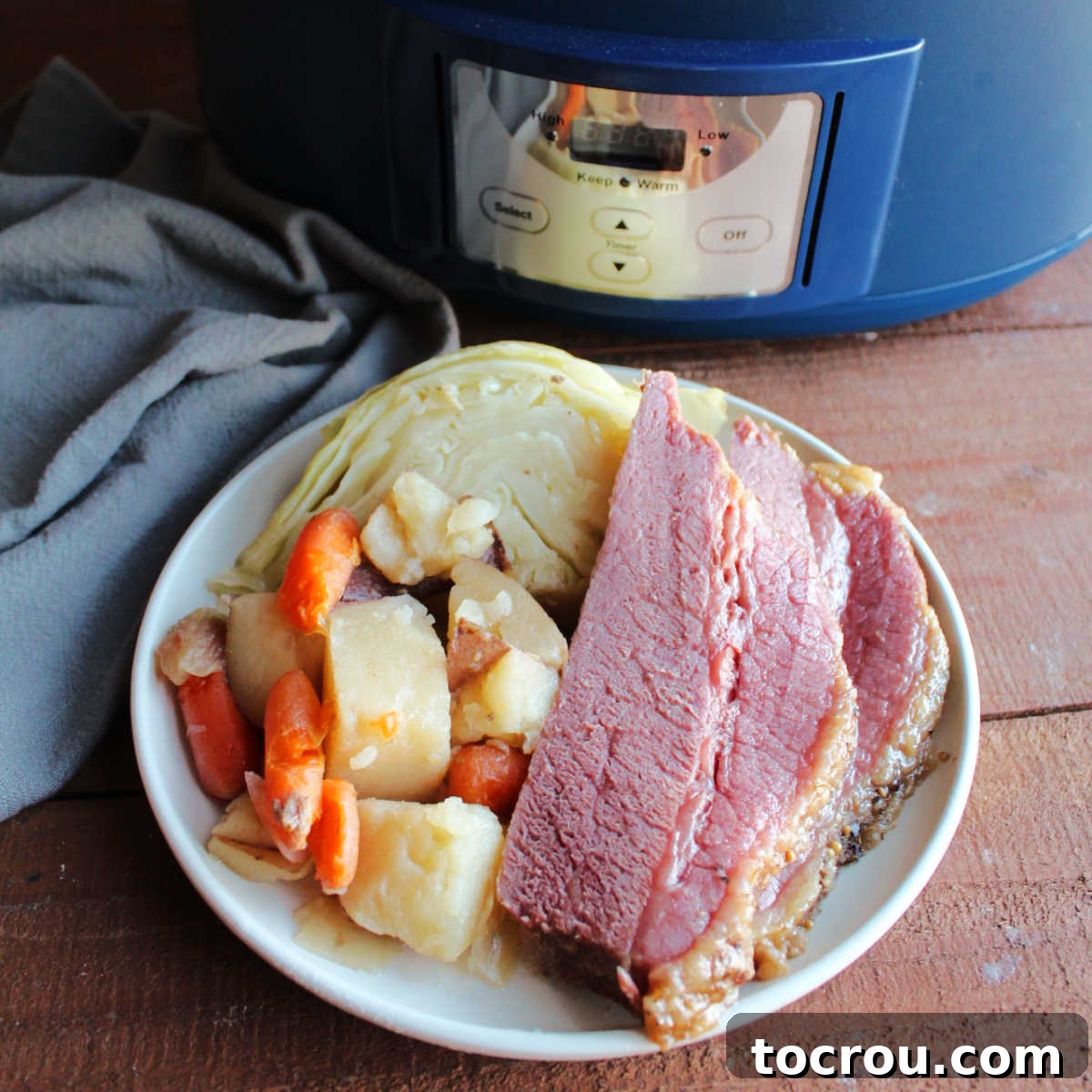 Plate of sliced corned beef with cooked potatoes, carrots, and cabbage wedge by the crockpot it was cooked in.