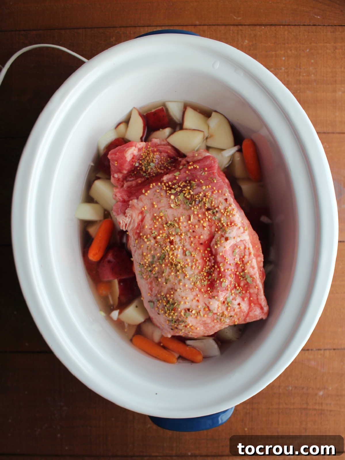 Chunk of corned beef on top of veggies in crockpot sprinkled with corned beef seasonings, ready to cook. 