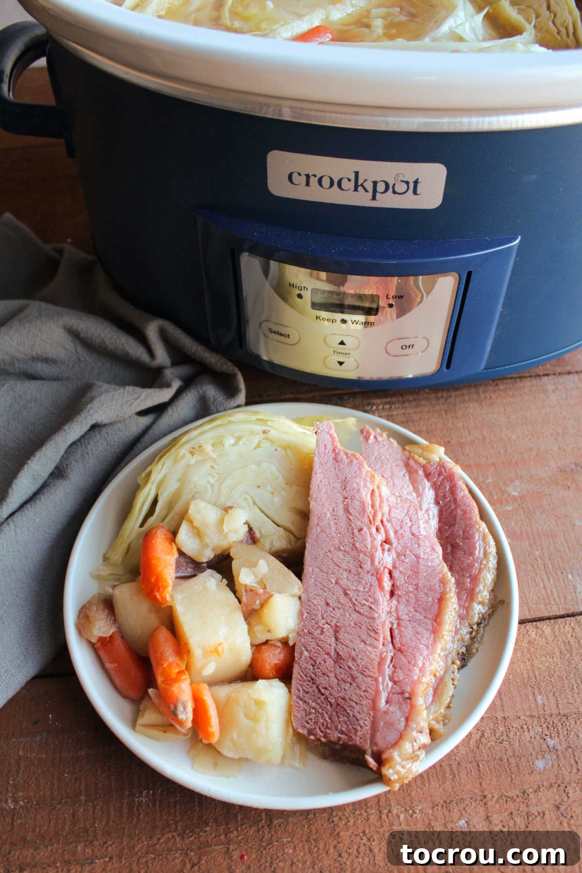 Plate with slow cooker corned beef, cabbage, potatoes, and carrots by the crockpot. 