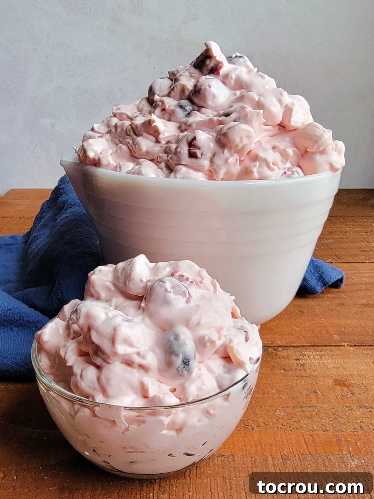 Looking across two bowls piled high with junk yard fruit salad with cherries, pineapple, blueberries, and more in fluffy pink dressing. 