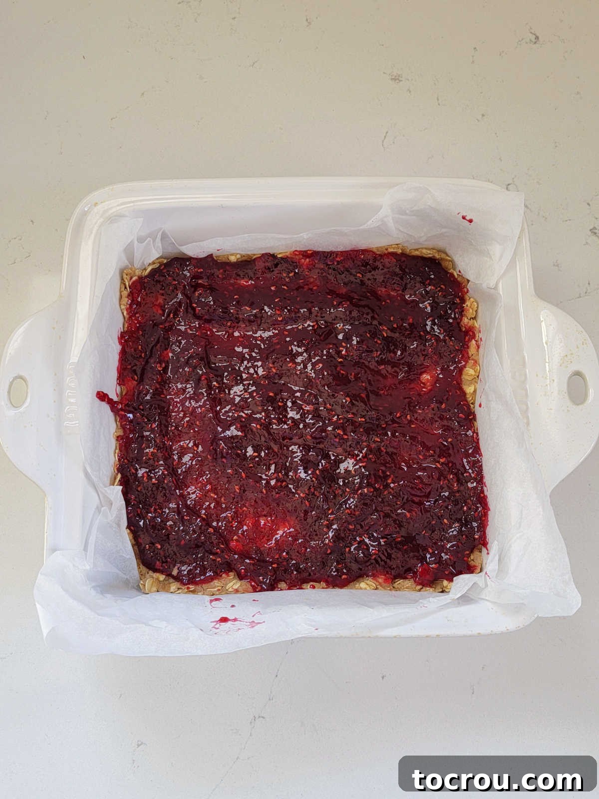 Raspberry jam evenly spread over the bottom crust of sourdough breakfast bars in a square baking pan.