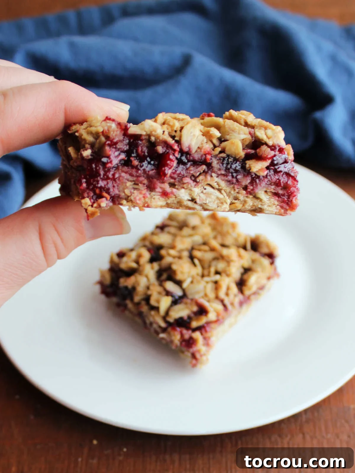 A hand holding a freshly baked sourdough breakfast bar, showing the crumbly oat topping and luscious raspberry jam filling.