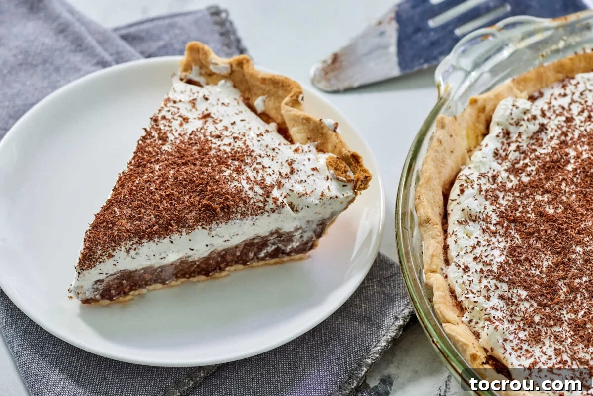 A beautifully crafted slice of homemade French silk pie, showcasing its rich, dark chocolate filling, a generous swirl of fluffy whipped cream, and delicate chocolate shavings on top. The remaining pie sits in the background, ready to be enjoyed.