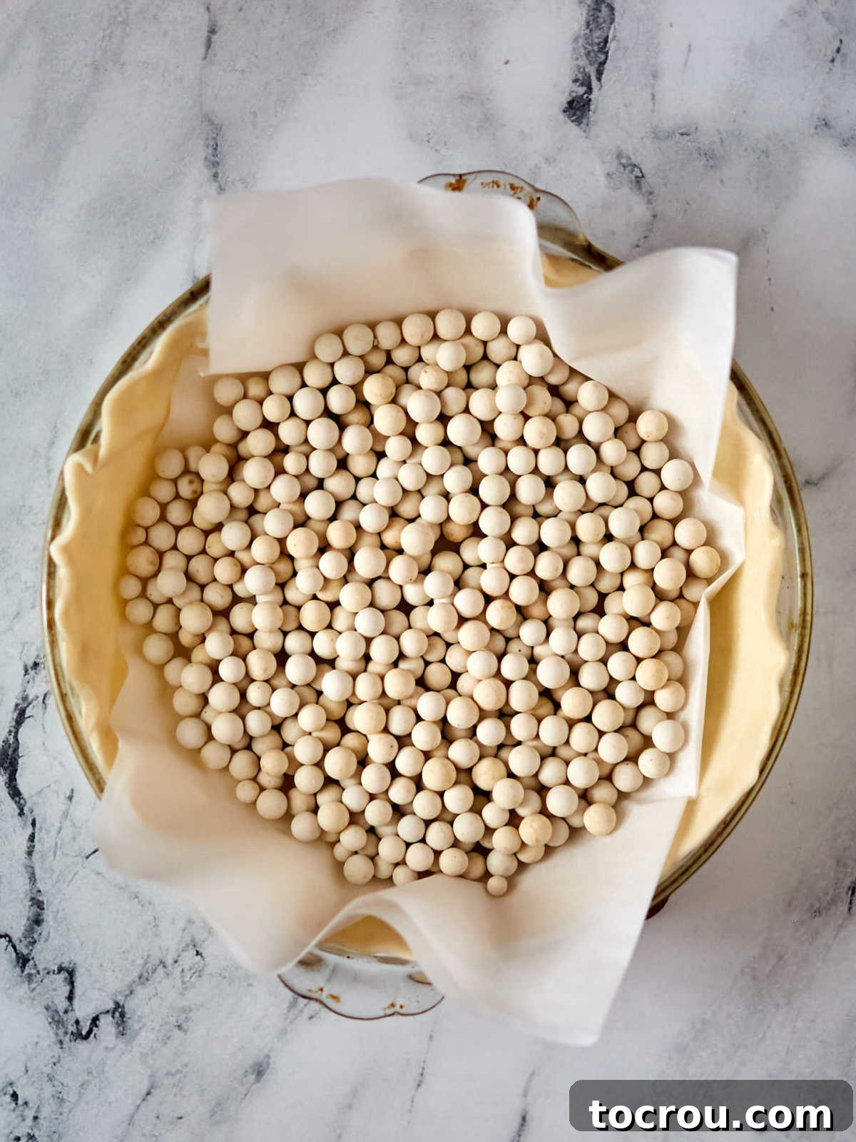 An unbaked pie crust expertly fitted into a glass pie plate, carefully lined with parchment paper and filled with ceramic pie weights, poised for blind baking to ensure a beautifully crisp and even base for the pie.