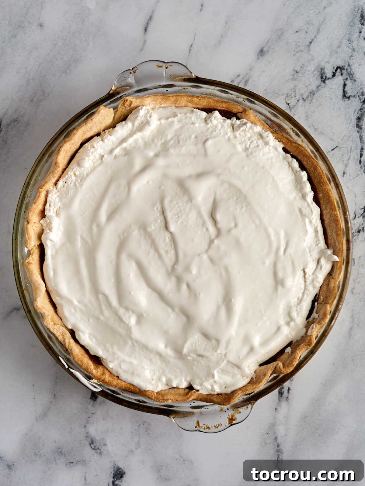 A fully assembled French silk pie, elegantly topped with a generous, swirling layer of fluffy, peak-forming whipped cream, patiently awaiting its final embellishment of delicate chocolate shavings.