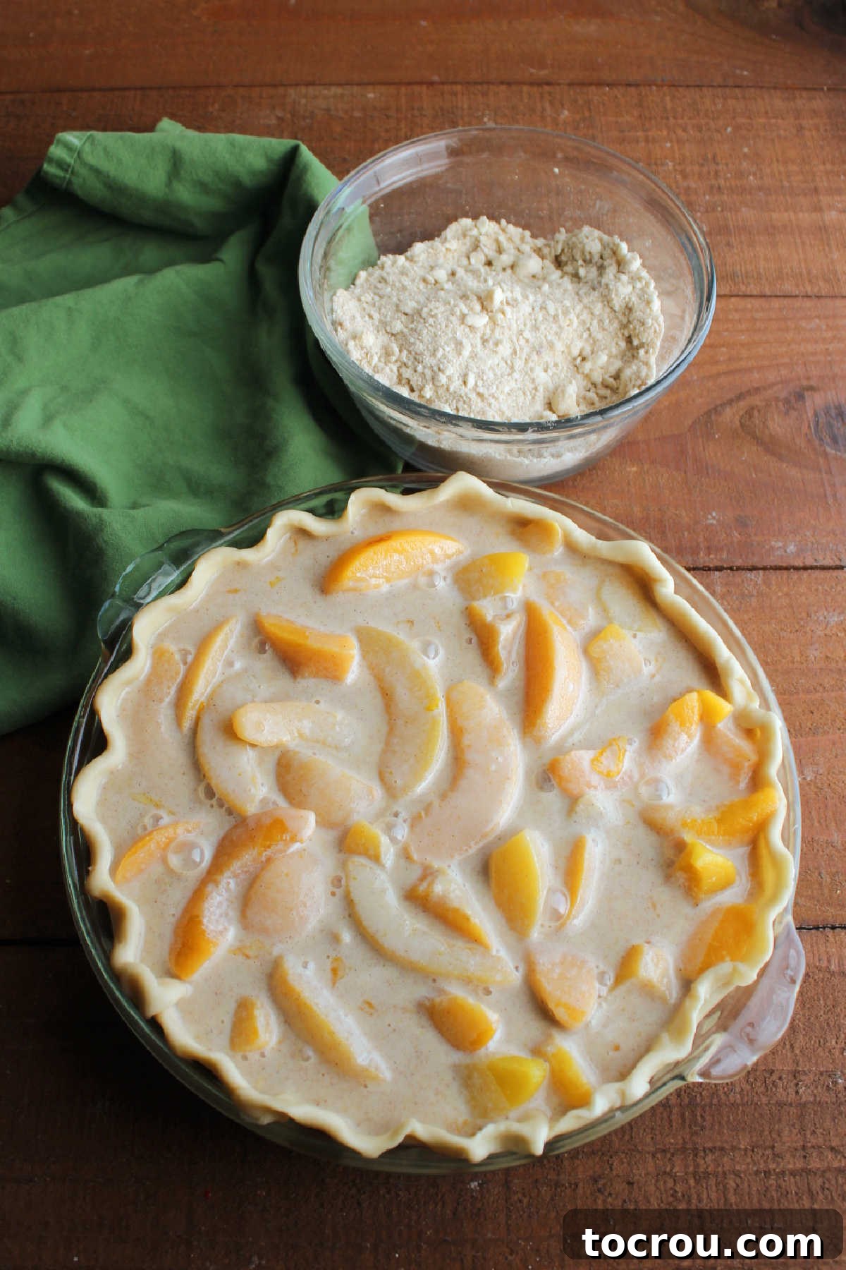 Pie with Filling and Streusel Ready Pie shell filled with peaches and custard mixture next to a small bowl with brown sugar streusel mixture.