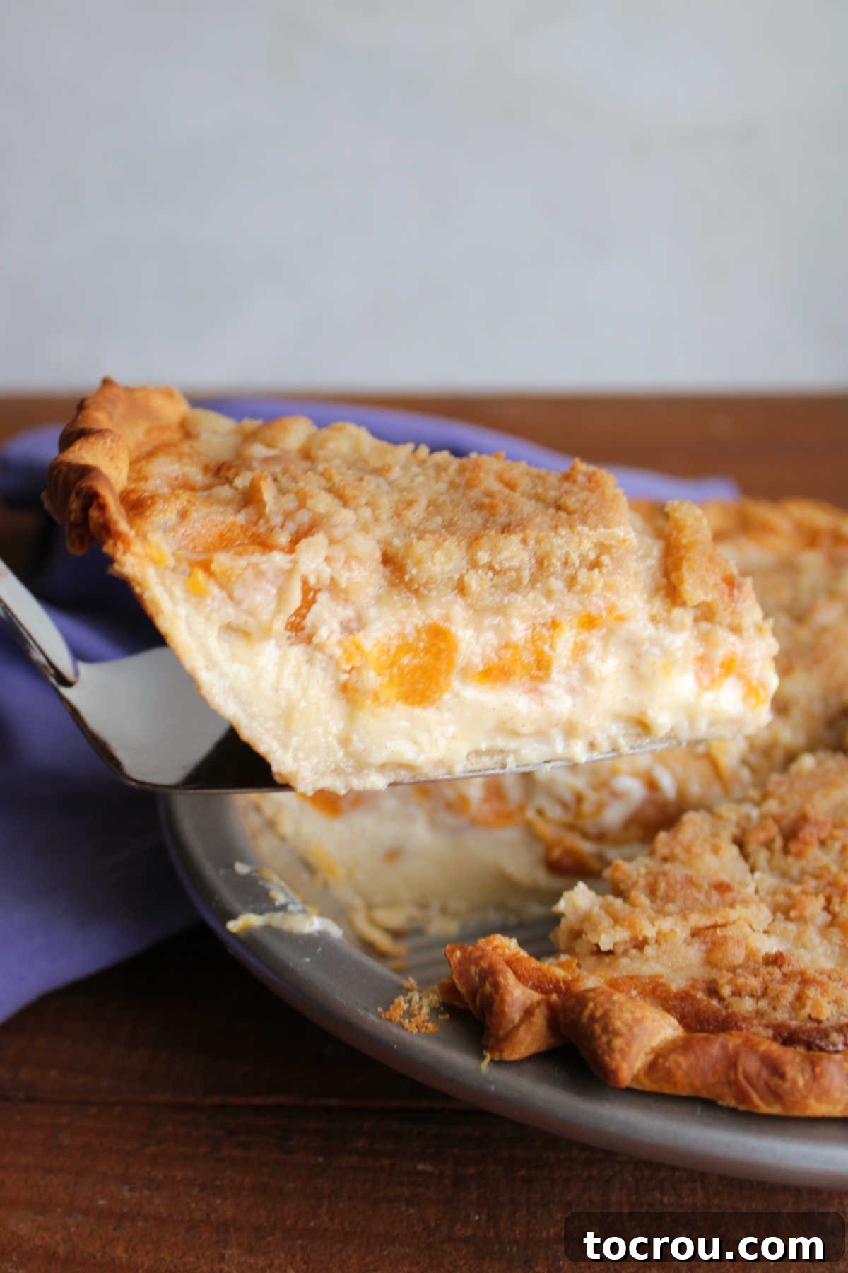Serving Peach Custard Pie Pie server lifting a piece of peach custard pie with chunks of peaches floating in creamy custard with streusel on top out of the pie pan.