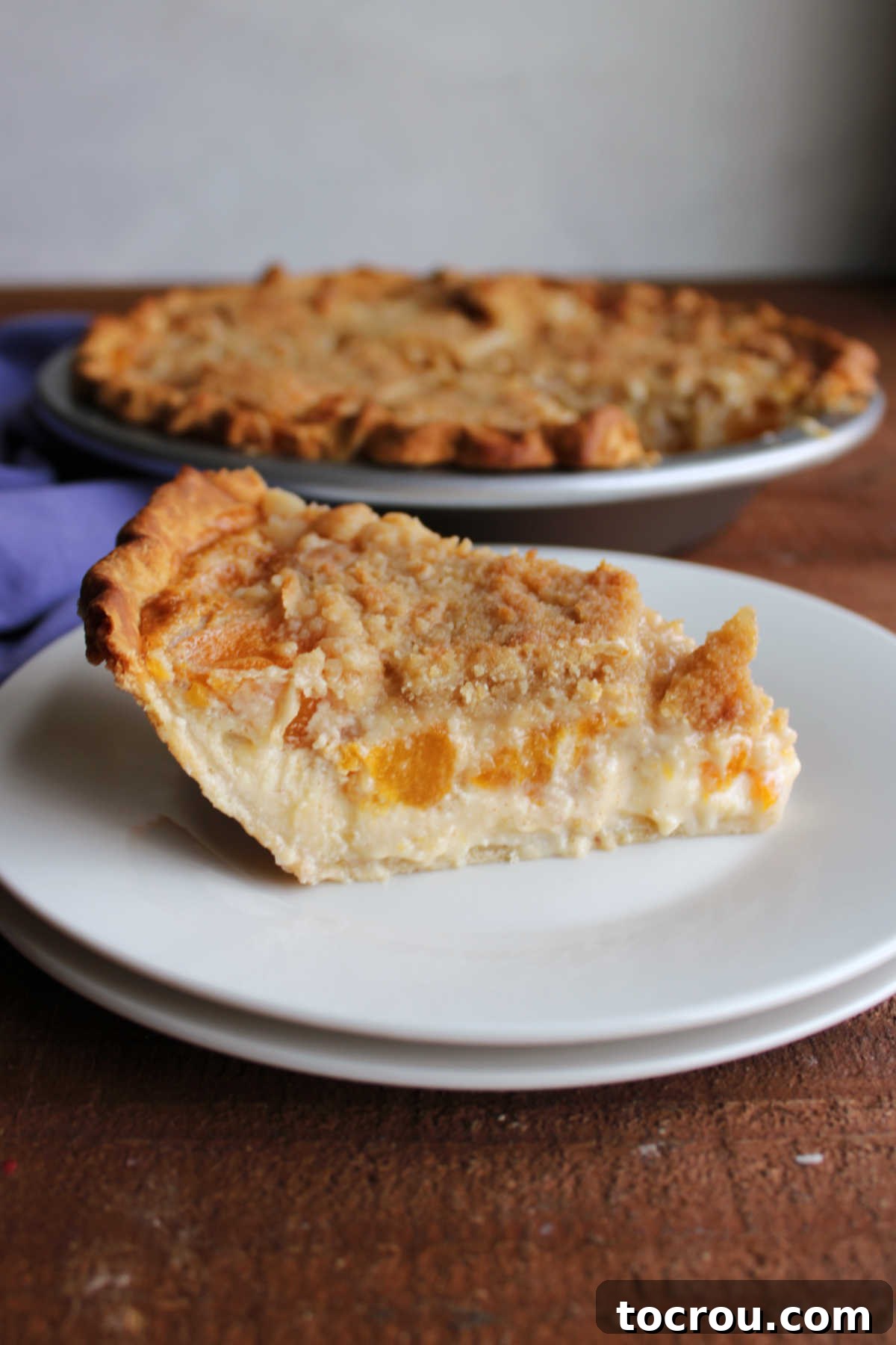 Individual Slice of Peach Custard Pie Slice of peach custard pie with crumb topping on dessert plate with remaining pie in background.