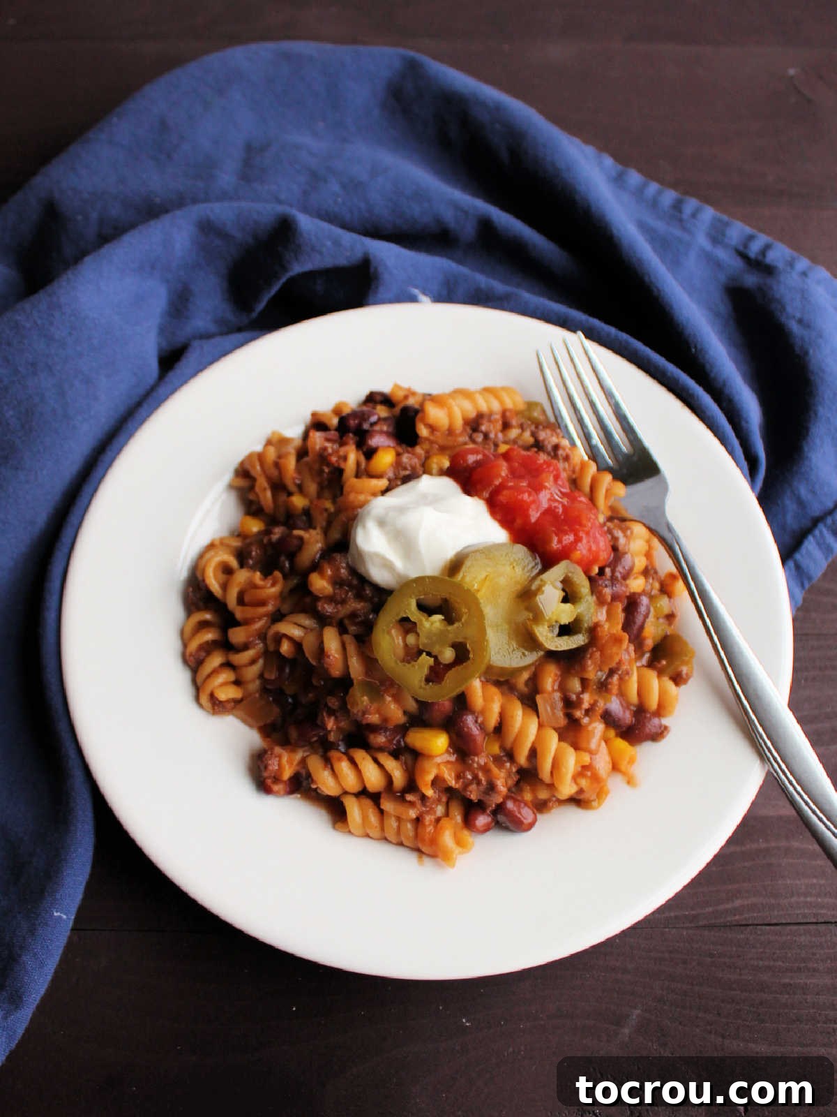 Plate of pasta with taco seasoning, ground beef, and veggies topped with sour cream, salsa, and pickled jalapenos.