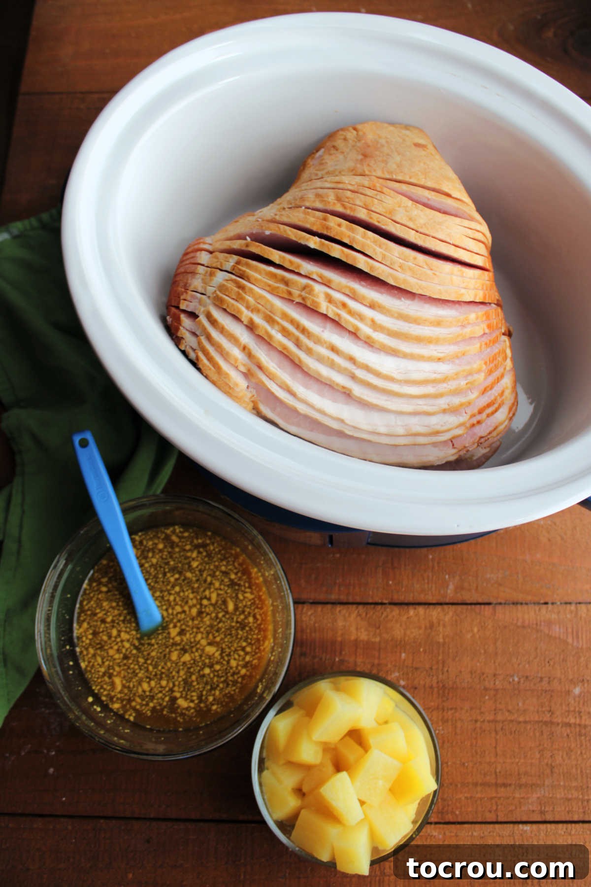 Golden Pineapple Glazed Slow Cooker Ham 3 Spiral cut ham in crockpot next to bowl of pineapple chunks and a bowl of pineapple juice, brown sugar, and mustard, ready to be put in crockpot to cook.