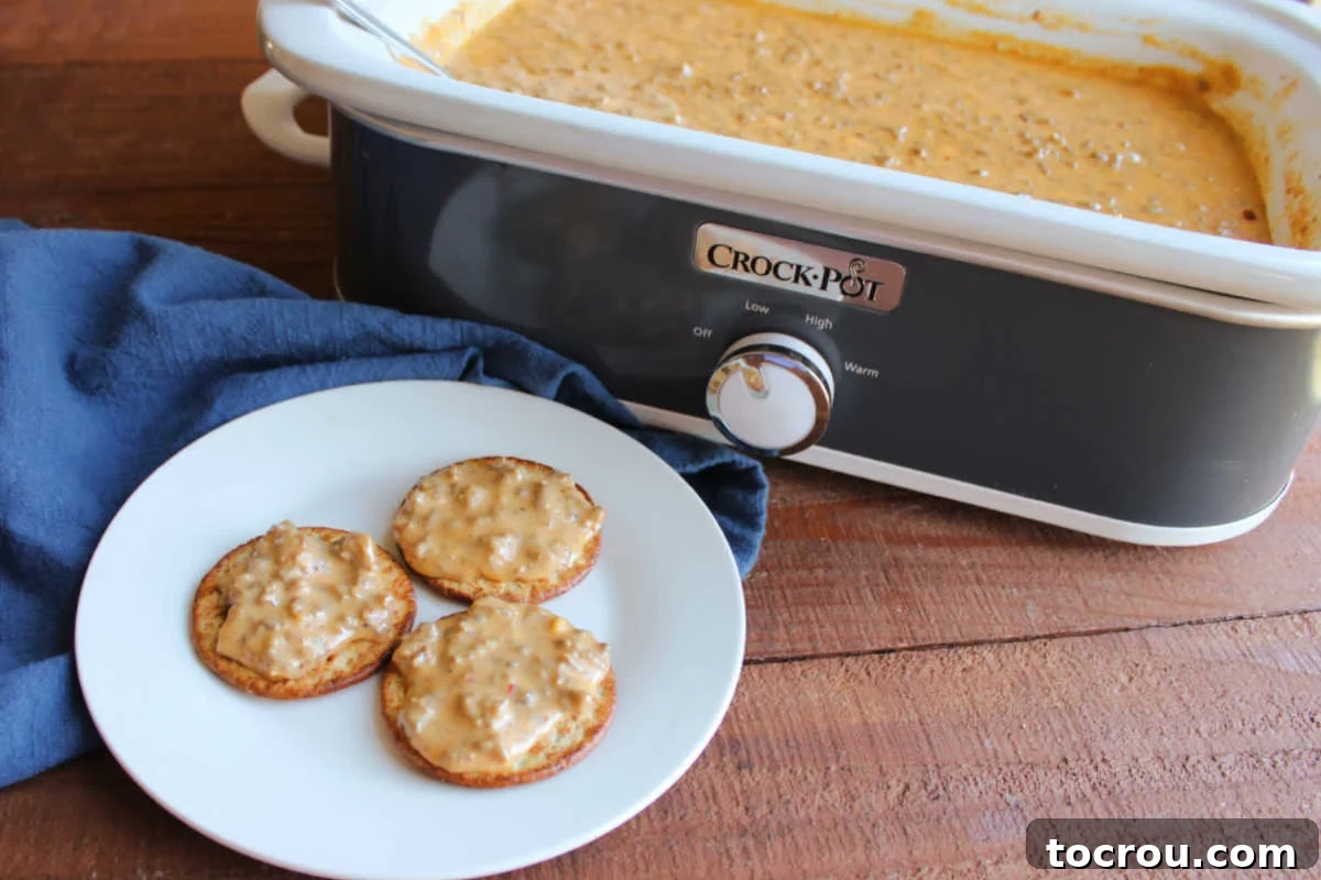 Crockpot filled with melted cheese dip with lots of sausage and hamburger in it next to a plate with crackers with some of the hanky panky dip on top.