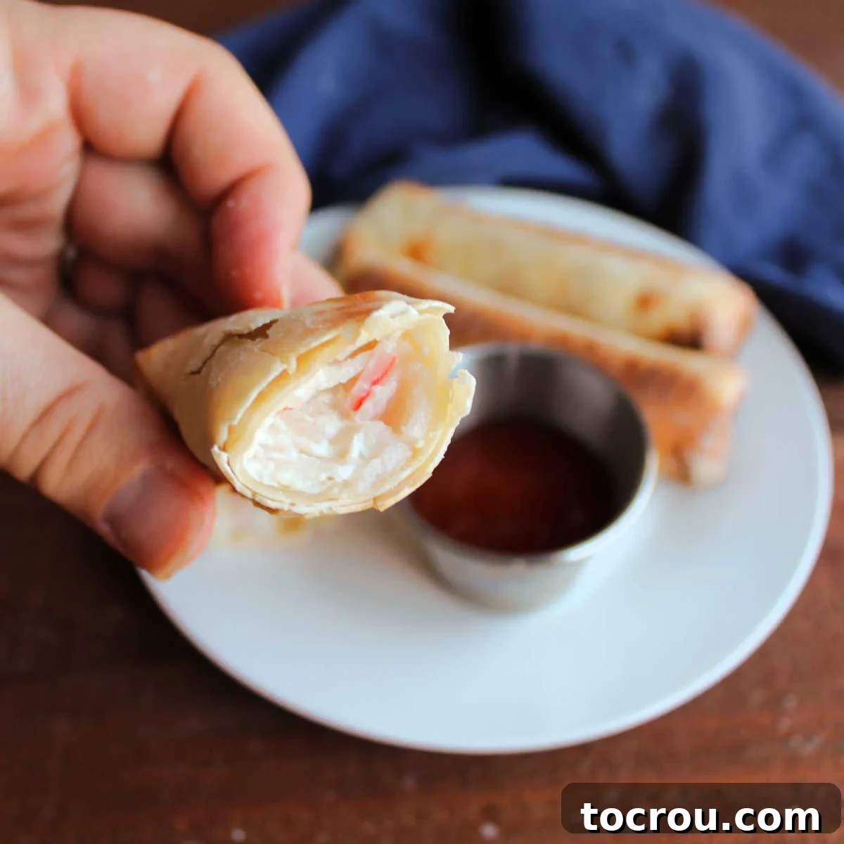 Baked Crab Rangoon Egg Roll with Creamy Filling Hand holding half of a baked crab rangoon egg roll, revealing the creamy filling with tender pink crab meat inside.