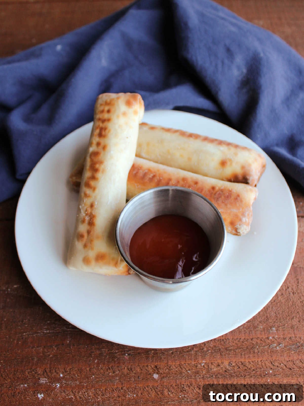Crab Rangoon Egg Rolls with Dipping Sauce A plate of golden brown baked crab rangoon egg rolls, beautifully arranged with a small container of sweet and sour sauce for dipping.