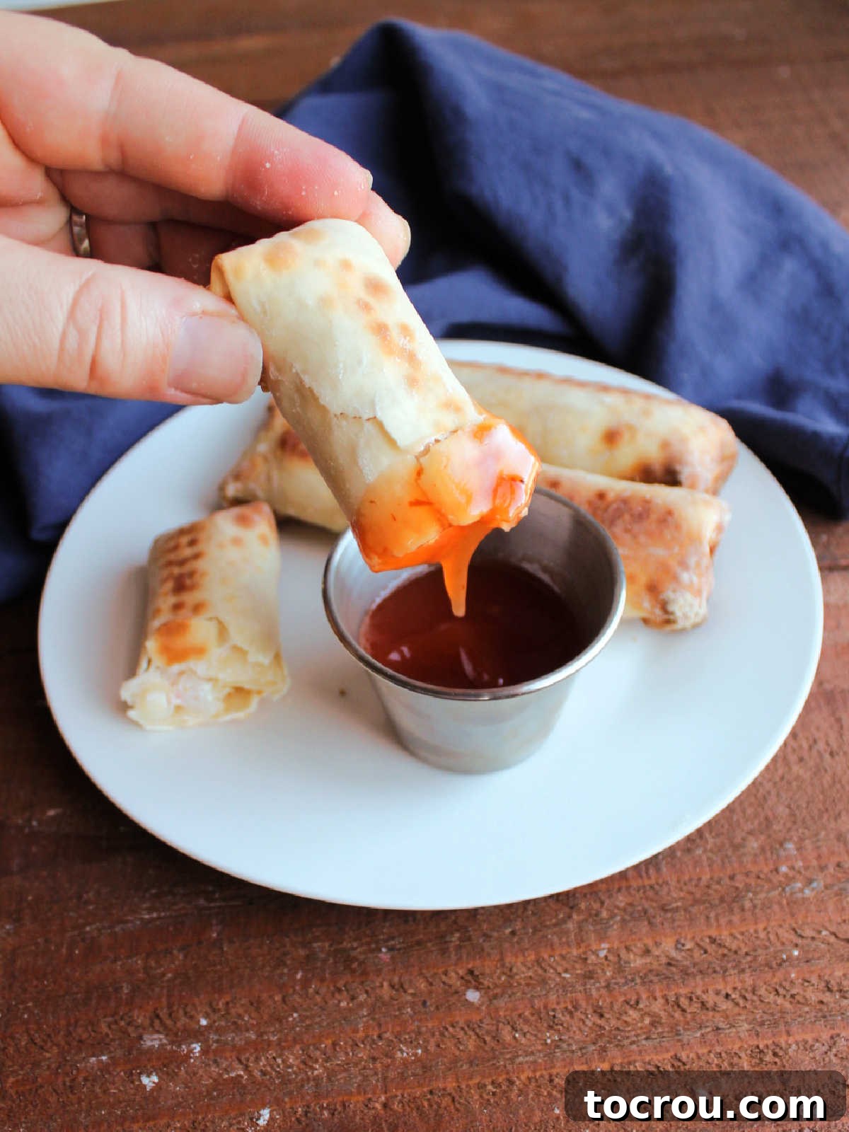 Dipping Crab Rangoon Egg Roll A hand dipping a crispy baked crab rangoon egg roll into a small bowl of sweet and sour sauce.
