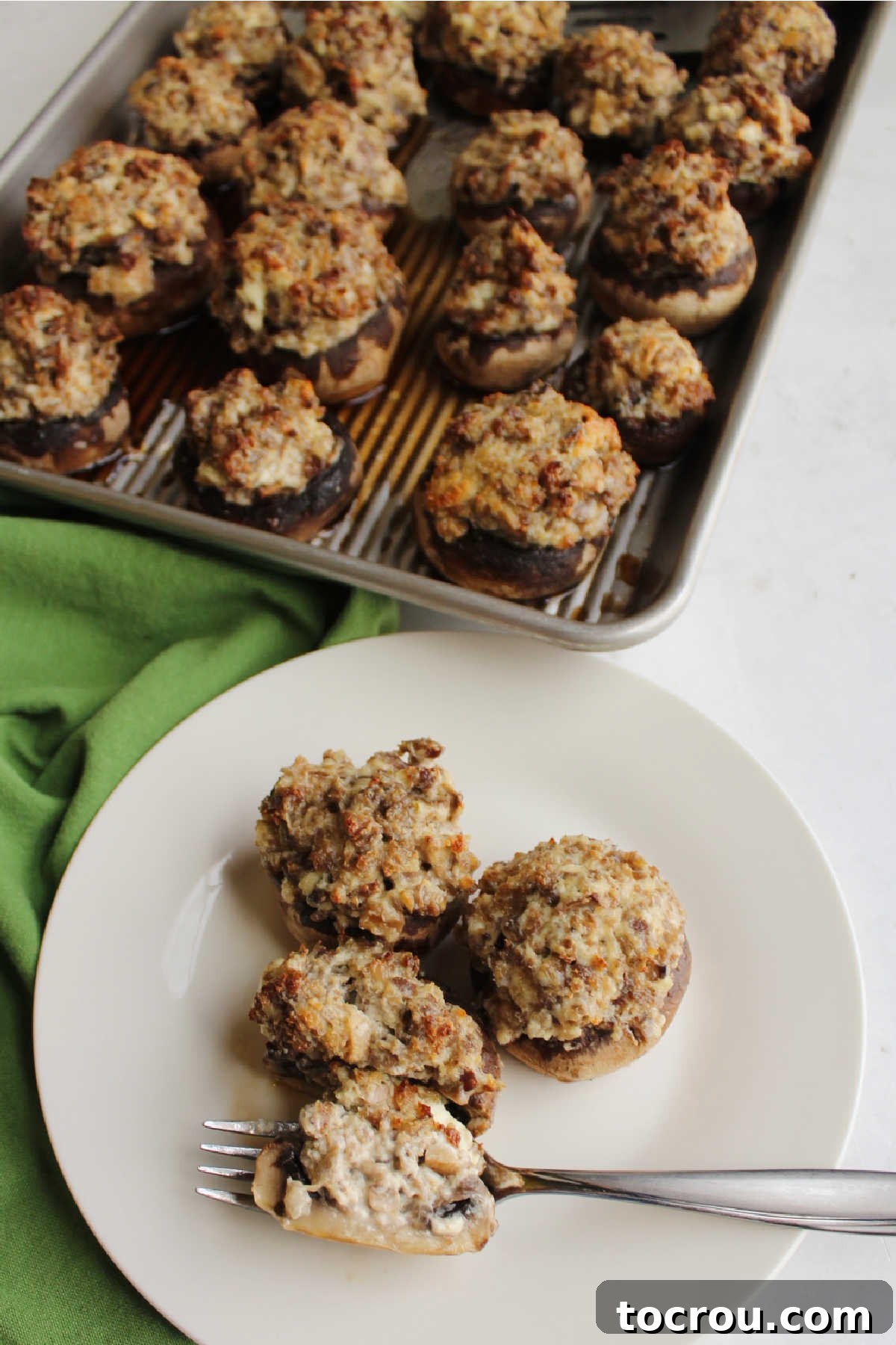 Savory Sausage Stuffed Mushrooms 8 Plate of sausage and cheese stuffed mushrooms with baking tray filled with remaining mushrooms in the background.