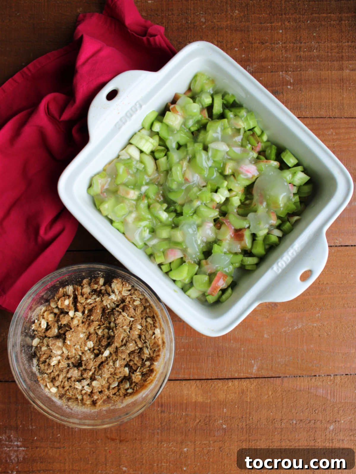 Square baking dish with sliced rhubarb and thickened sugar inside next to bowl of brown sugar oat topping. 