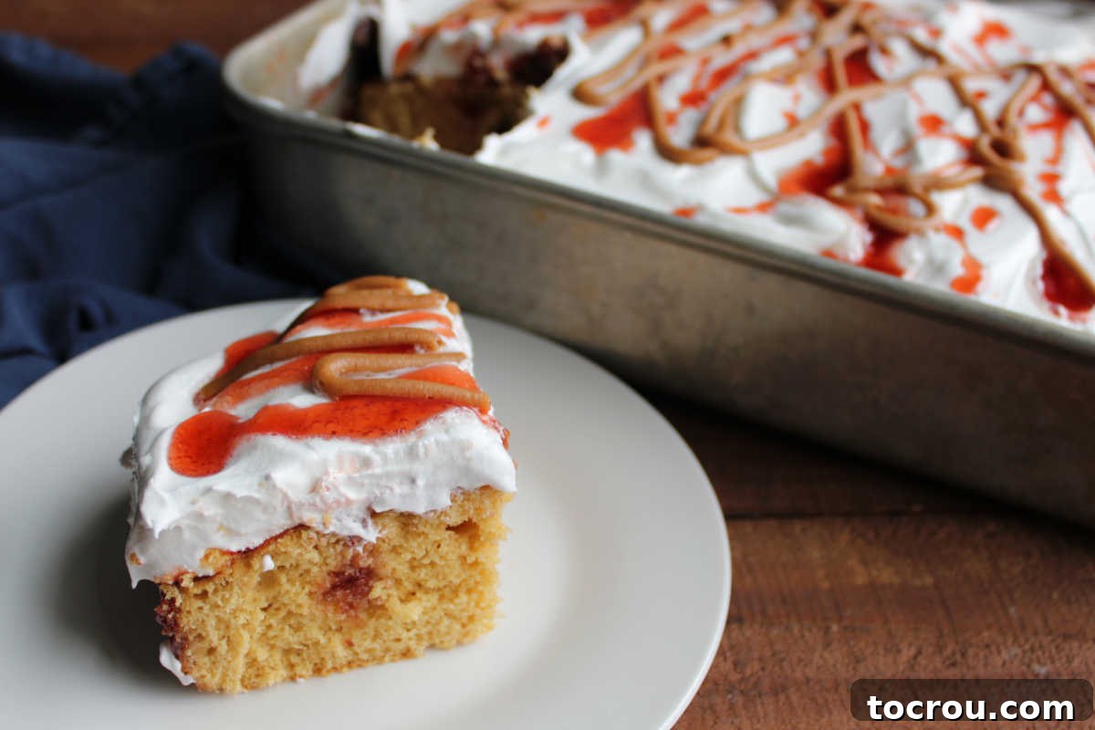 Classic Peanut Butter and Jelly Poke Cake Slice A delectable slice of peanut butter and jelly poke cake served on a plate, showcasing layers of moist peanut butter cake, vibrant jelly tunnels, and a fluffy whipped topping. The remaining cake sits in the pan in the background, ready to be enjoyed.