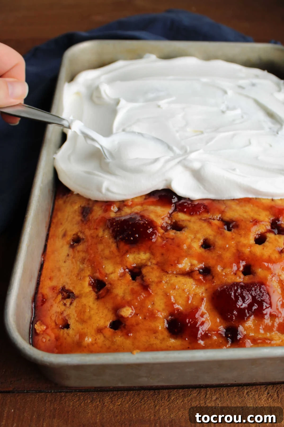 Applying Cool Whip Topping on Jelly Poke Cake Smooth, white Cool Whip being evenly spread over the cooled peanut butter poke cake, which is already infused with glistening strawberry jelly, creating a creamy top layer.