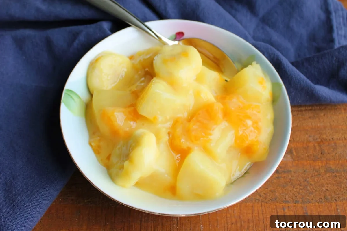 Small bowl of sunshine salad with pineapple chunks, oranges, and bananas in creamy vanilla pudding dressing with spoon, ready to eat.