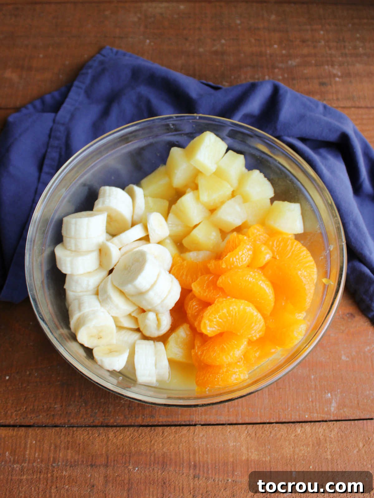 Big glass mixing bowl with sliced banana, mandarin oranges, and pineapple chunks inside.