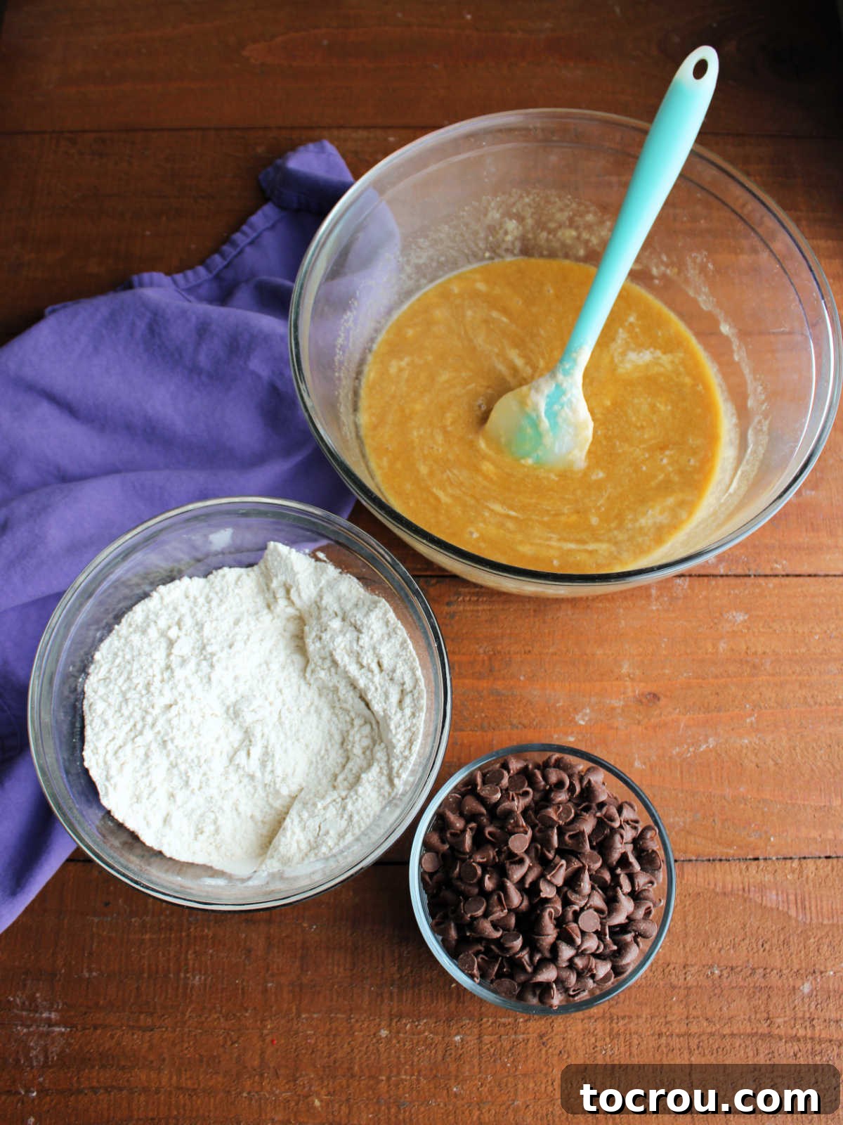 A large mixing bowl containing melted butter, brown sugar, granulated sugar, and sourdough discard, ready for combining. Adjacent are a smaller bowl of dry ingredients and a separate bowl brimming with chocolate chips.