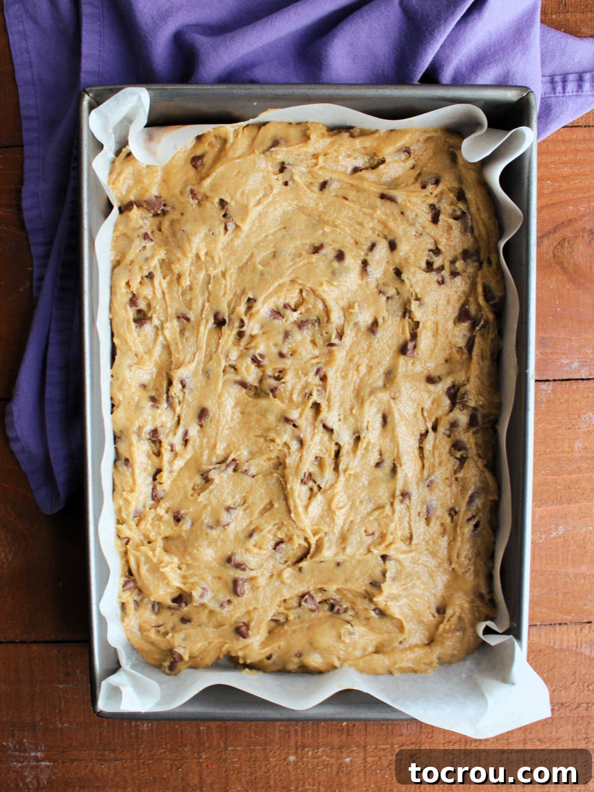 Sourdough chocolate chip cookie dough evenly pressed into a parchment-lined 9x13-inch baking pan, ready for the oven.