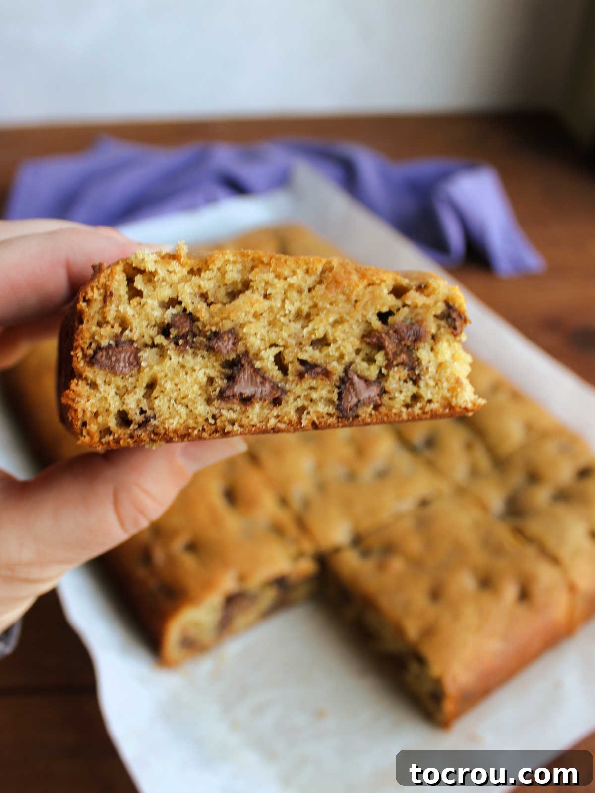 A hand holds a freshly cut sourdough chocolate chip cookie bar, showcasing its soft, moist interior generously studded with melted chocolate chips.