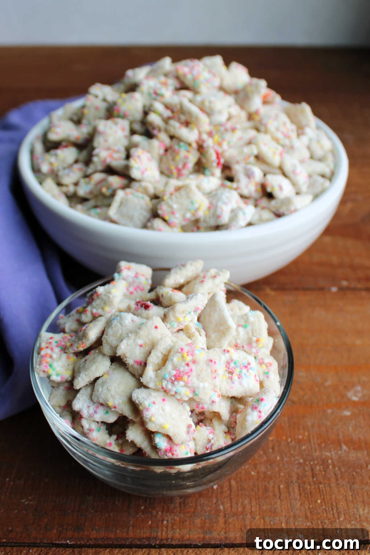 Small bowl filled with birthday cake puppy chow with more in a serving bowl behind it showing creamy white coating with lots of colorful sprinkles. 