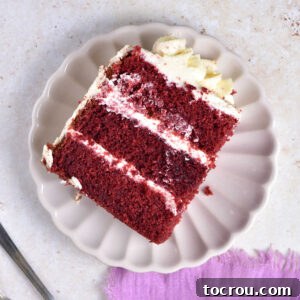 Piece of 3 layer red velvet cake with creamy frosting on plate, ready to eat.