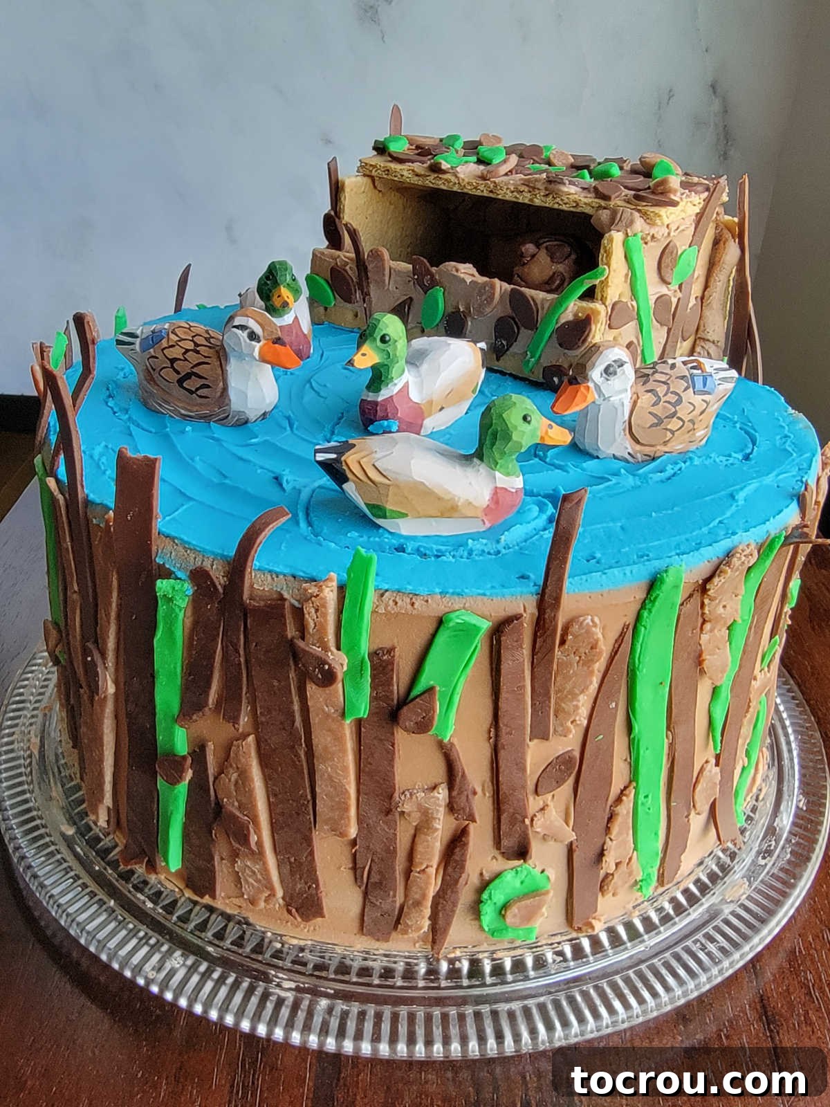 Duck Hunting Themed Cake with Modeling Chocolate Decor Duck hunting cake with modeling chocolate strips decorating the side like camouflage, modeling chocolate leaves on graham cracker blind, and modeling chocolate figurine inside.