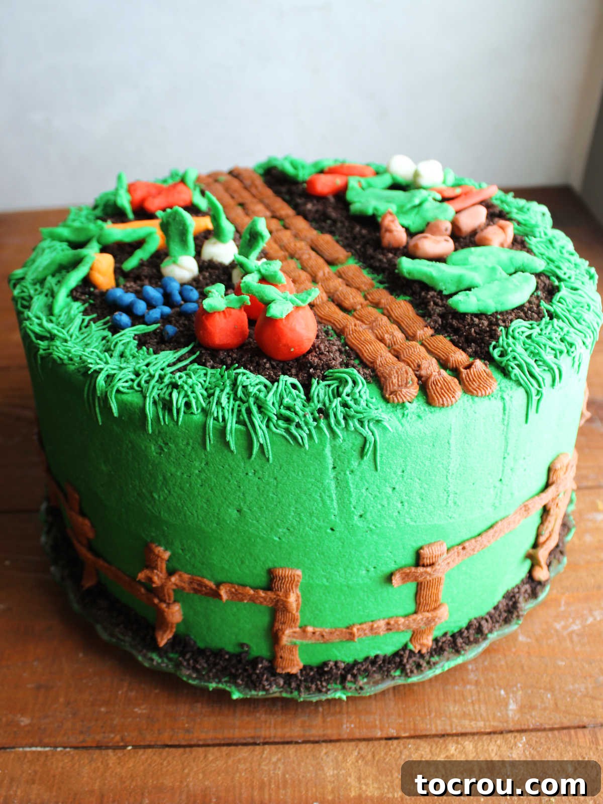 Garden Themed Cake Adorned with Modeling Chocolate Produce Garden themed cake with green frosting and brown frosting fence on the outside with Oreo crumb dirt on top covered with modeling chocolate fruits and vegetables like tomatoes, pea pods, carrots, strawberries, onions, blueberries, etc.