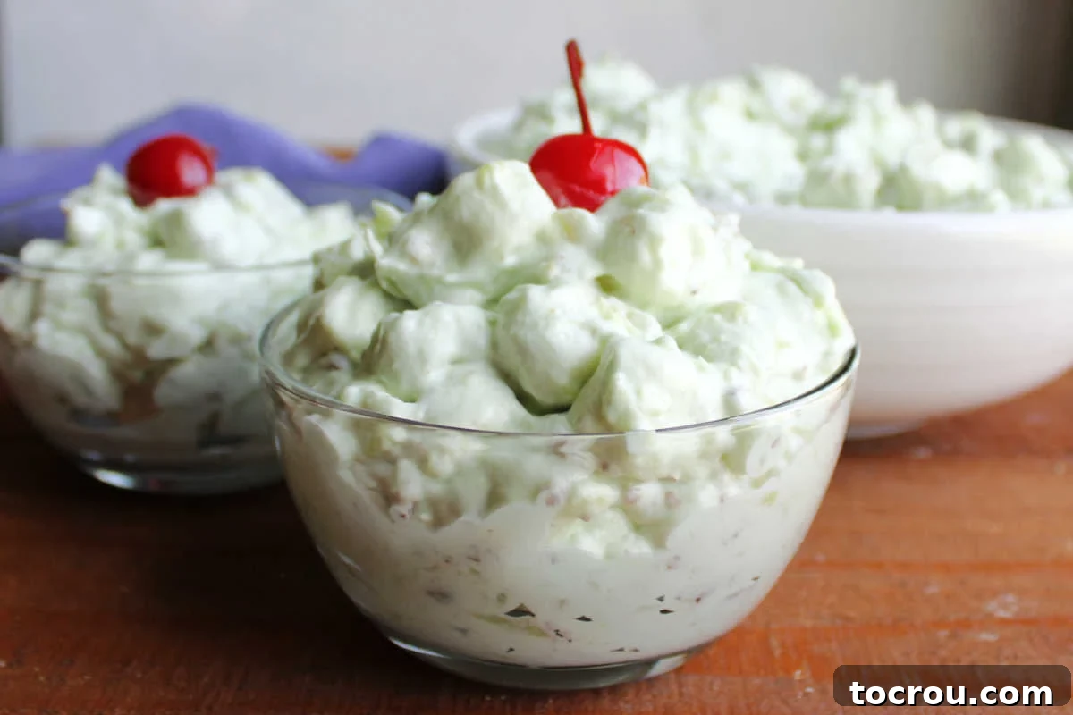 Two small bowls filled with fluffy watergate salad topped with maraschino cherries with serving bowl of more pistachio fluff in the background. 