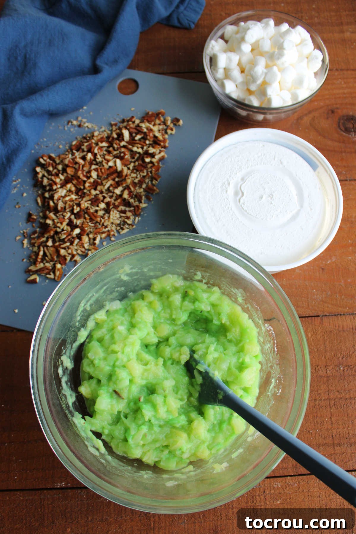 Making watergate salad with mixing bowl filled with pudding mix and crushed pineapple with remaining ingredients nearby. 