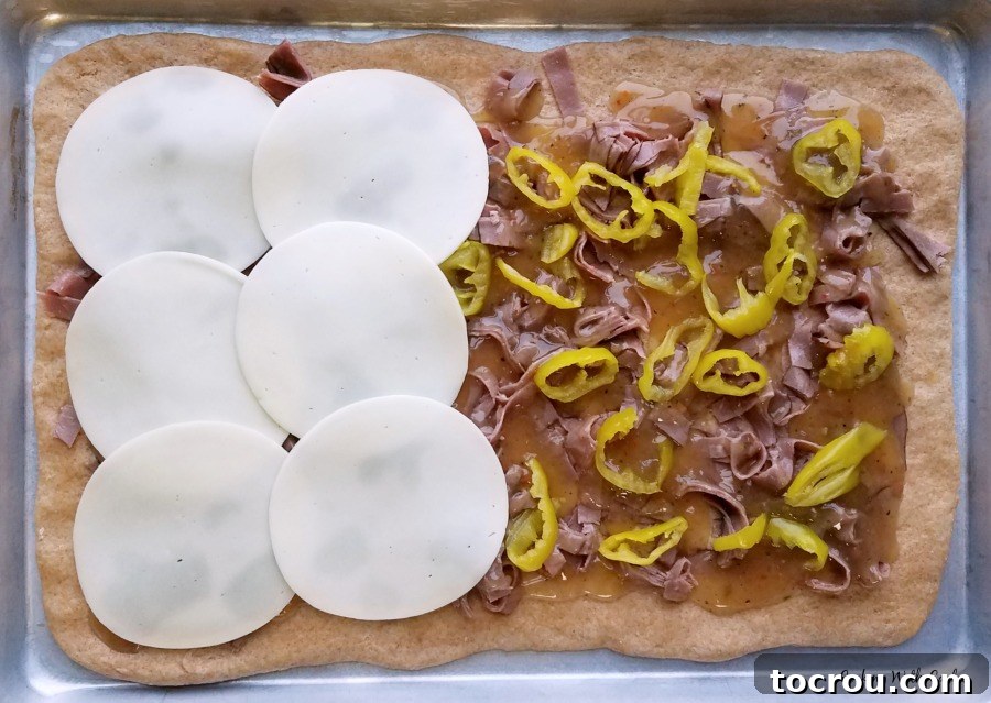 Half sheet pan filled with pizza crust topped with sliced beef, pepperonicini rings and a layer of provolone going over the top.