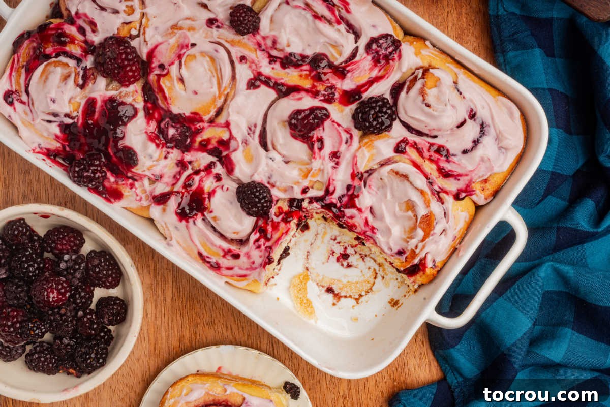 Baking pan filled with blackberry sweet rolls topped with fruity cream cheese frosting and blackberry compote with one roll missing.