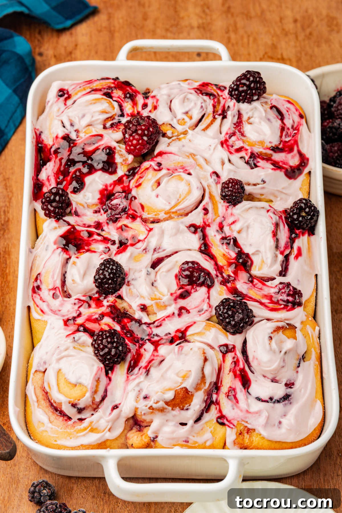Baking pan filled with finished blackberry sweet rolls showing swirls of blackberry compote in the fluffy rolls topped with cream cheese frosting and more blackberry compote.