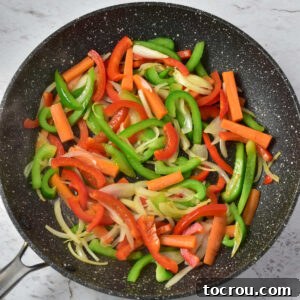 Skillet with strips of bell pepper, onions, and carrots cooking in hot oil.