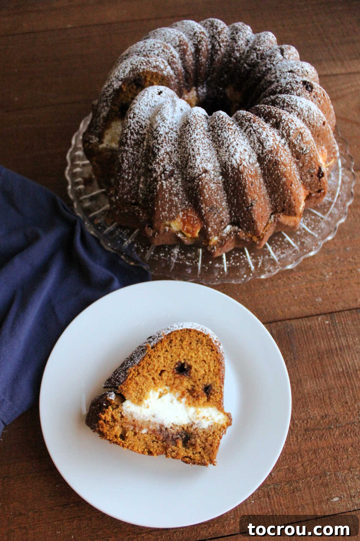 Harvest Pumpkin Bundt Cake with Cream Cheese Core 9 A beautiful slice of cream cheese-stuffed pumpkin Bundt cake with delightful chocolate chips, presented on a plate, with the rest of the powdered sugar-dusted cake visible in the background.