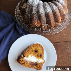 Harvest Pumpkin Bundt Cake with Cream Cheese Core 10 Slice of pumpkin bundt cake with cream cheese filling and chocolate chips on plate with remaining powdered sugar dusted cake in background.