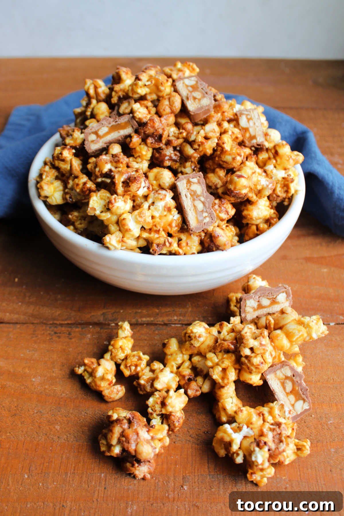 Serving bowl spilling over with homemade snickers popcorn feature chocolate, caramel, peanuts, and candy bar in crunchy popcorn. 