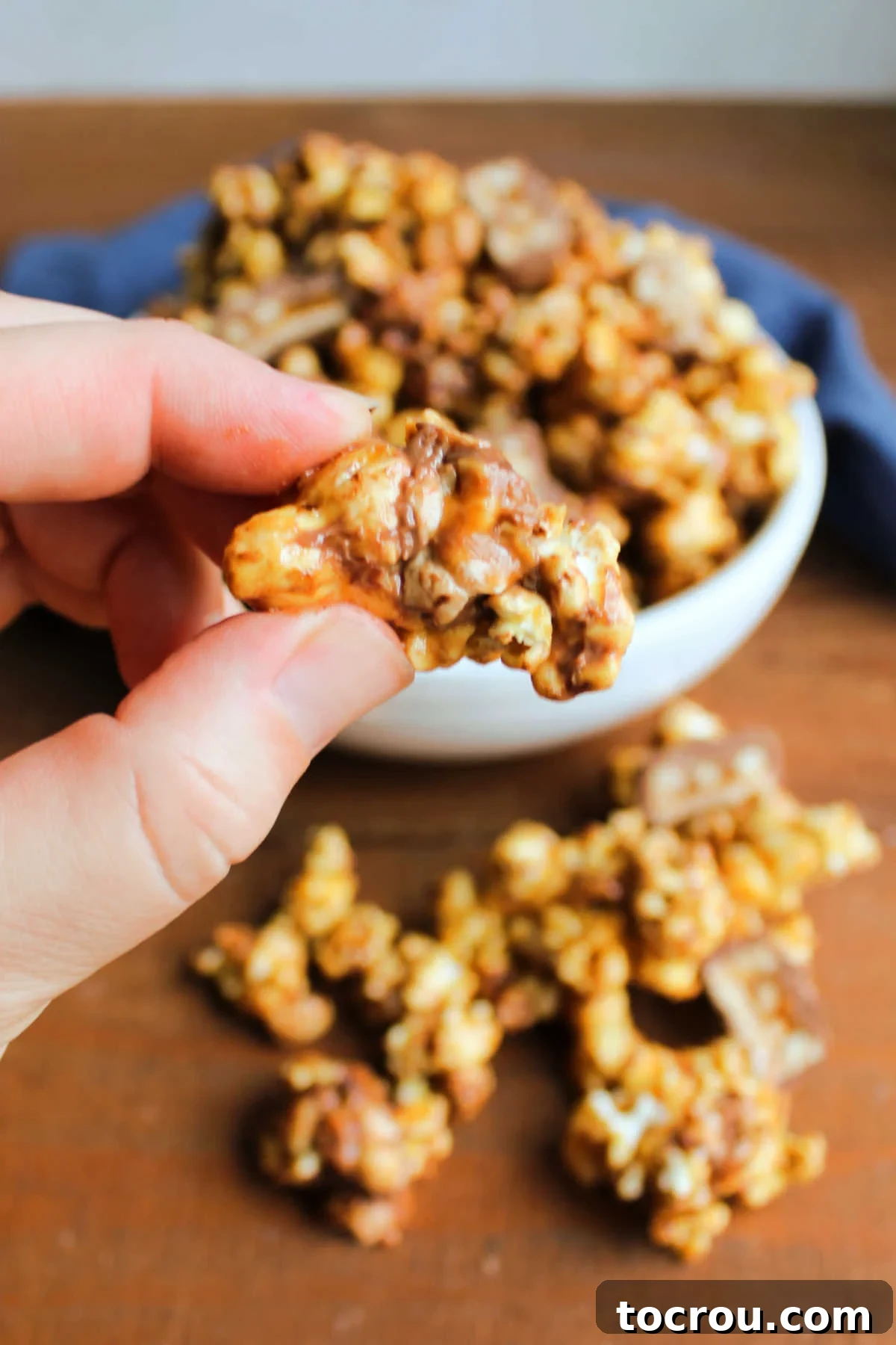 Hand holding a cluster of snickers popcorn showing the crunchy caramel coating with peanuts, popcorn, chocolate and candy bits. 