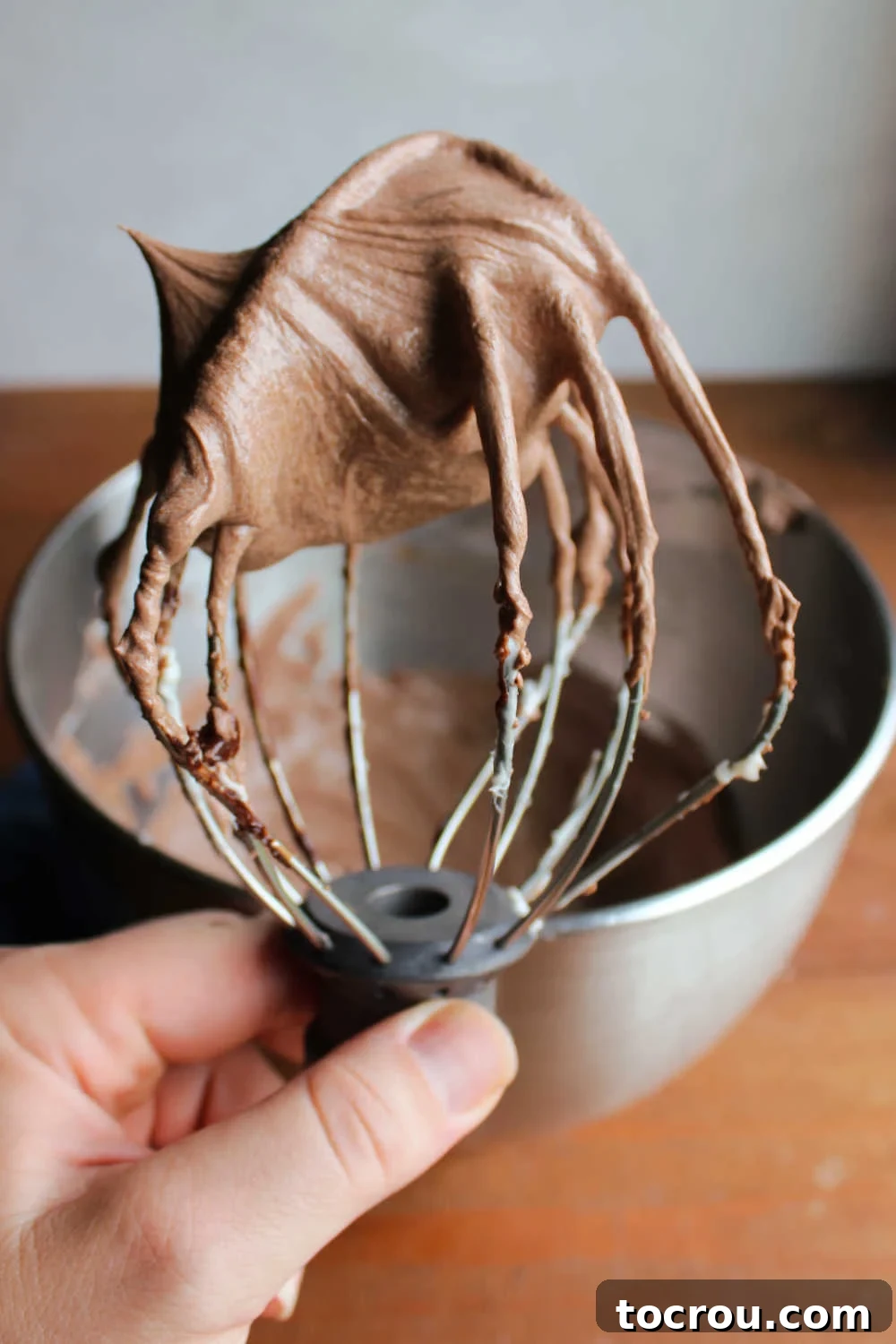 Hand holding whisk attachment with fluffy chocolate ermine frosting on it, with a mixer bowl full of remaining frosting in the soft-focus background.