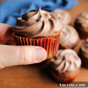 Hand holding a chocolate pumpkin cupcake topped with a swirl of chocolate ermine frosting, perfectly piped.