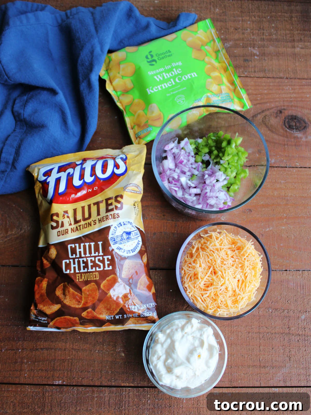 Zesty Frito Corn Crunch 3 Ingredients for Frito corn salad, including chili cheese Fritos, frozen corn, red onion, bell pepper, shredded cheddar cheese, and mayonnaise, laid out and ready to be combined.