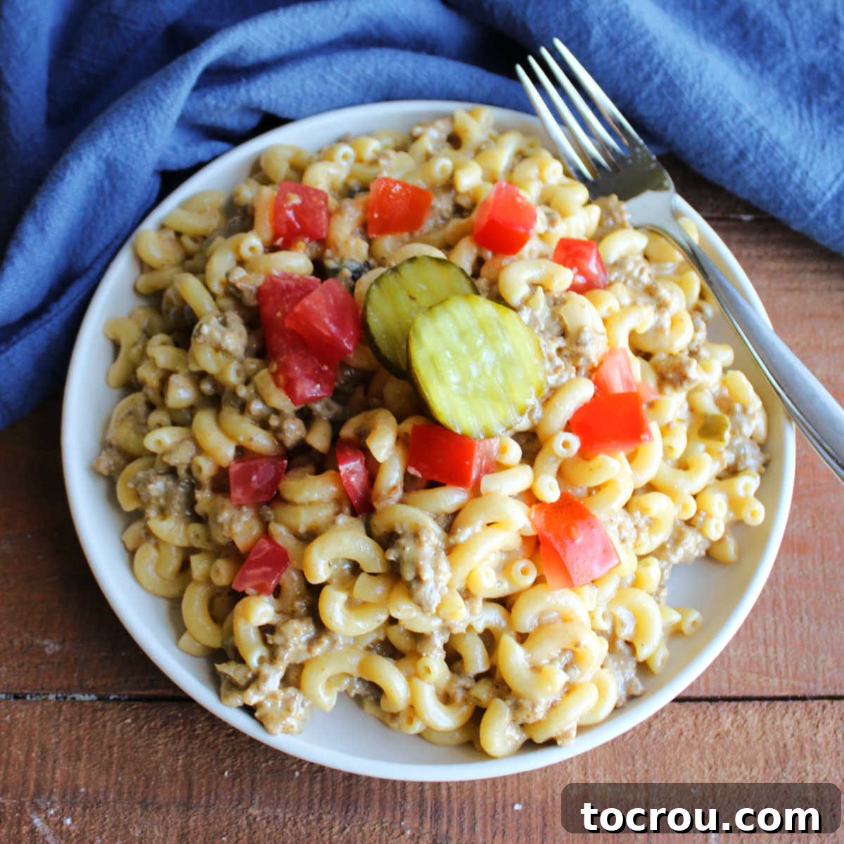 A vibrant plate of cheeseburger pasta, generously topped with fresh diced tomatoes and a scattering of crisp bread and butter pickle chips, ready to be enjoyed.