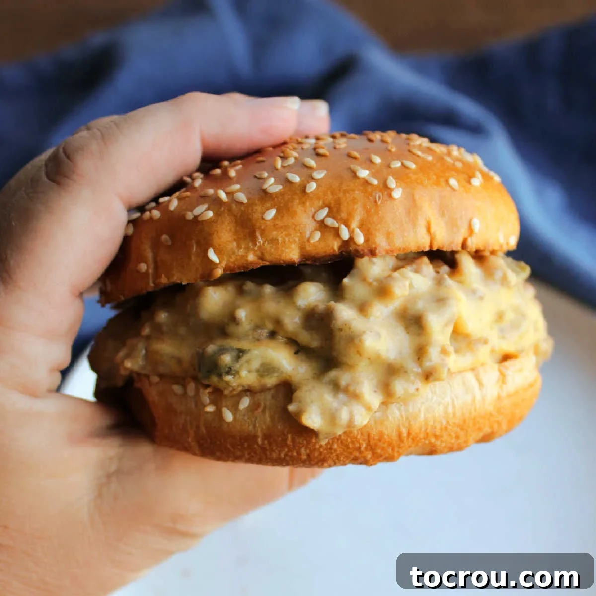 Hand holding a toasted sesame bun filled with crockpot cheeseburger mixture, showcasing the rich, cheesy filling.