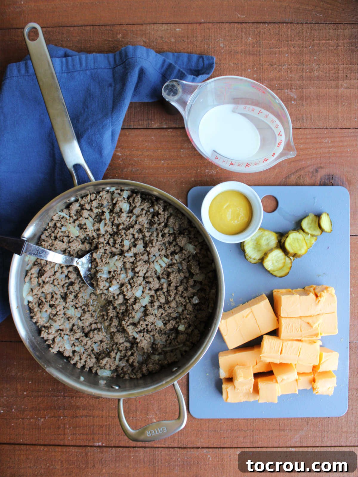 Ingredients laid out for crockpot cheeseburgers, including ground beef, diced onions, cubed Velveeta cheese, bread and butter pickles, and yellow mustard.