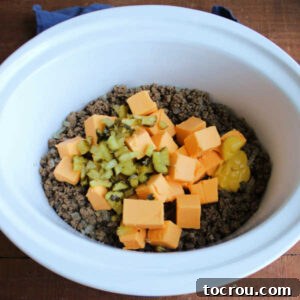 Browned ground beef mixture in crockpot topped with cubes of Velveeta cheese, pickles, and mustard, ready for slow cooking.