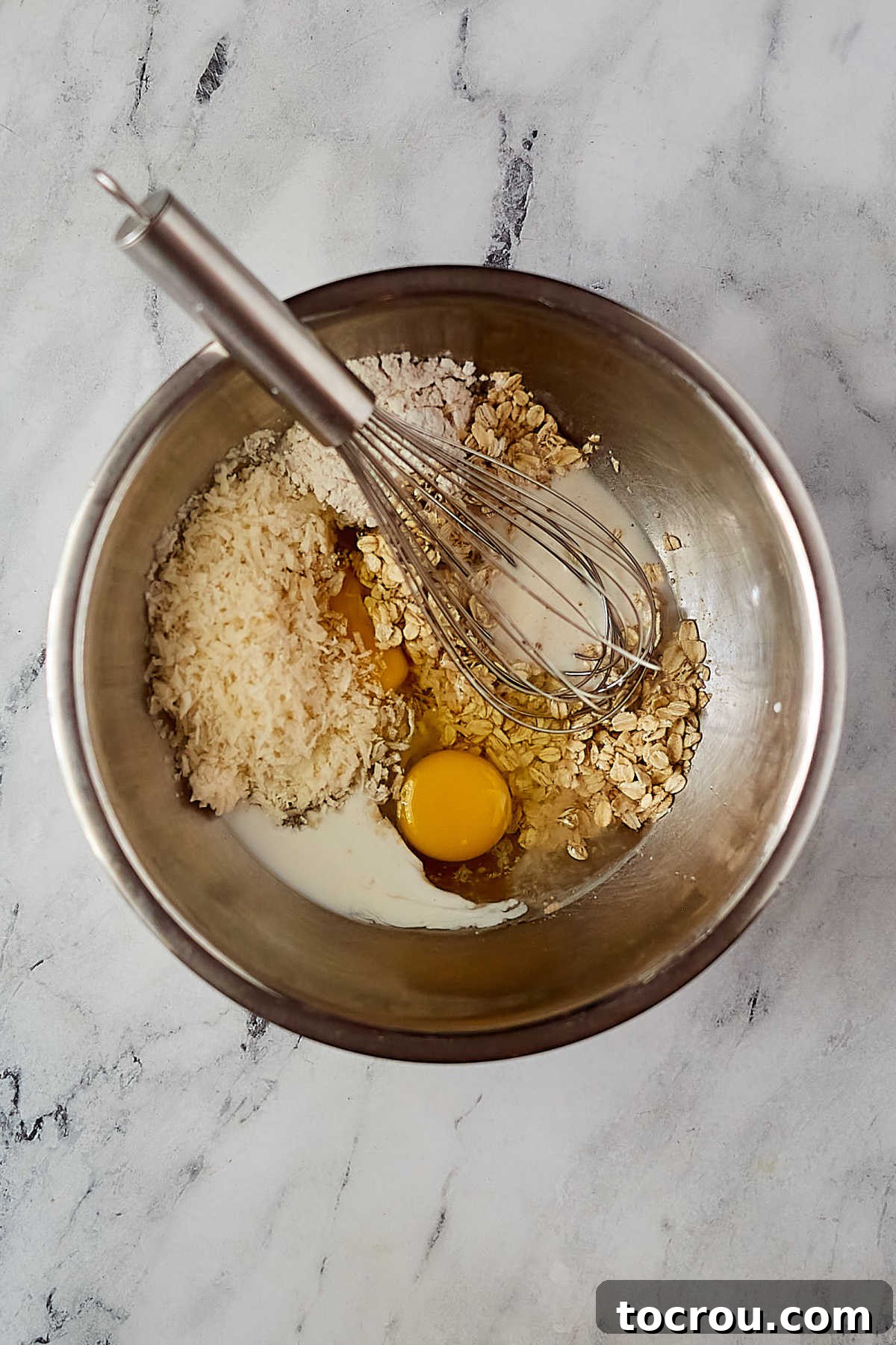 A large mixing bowl containing the wet and dry ingredients for oatmeal pie filling – eggs, corn syrup, milk, melted butter, vanilla, salt, flour, coconut, and oats – ready to be gently stirred together before pouring into the pie crust.