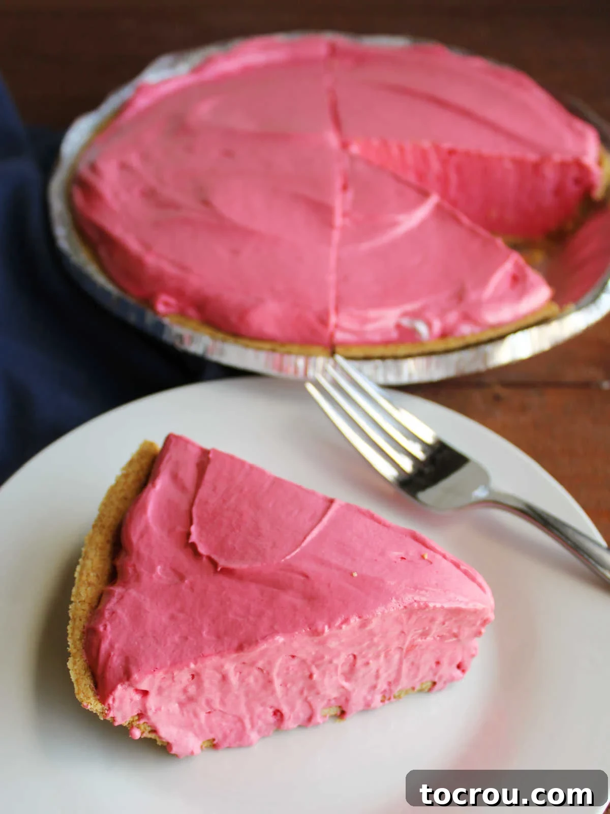 Slice of fruit punch Kool-Aid pie with graham cracker crust on plate with fork and remaining pie nearby. 
